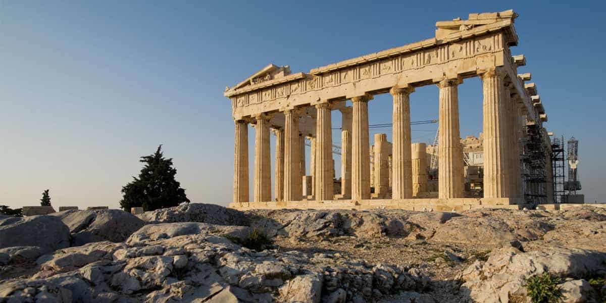 The Parthenon, an ancient Greek temple with tall columns, stands atop a rocky hill under a clear blue sky, partially surrounded by scaffolding for restoration.
