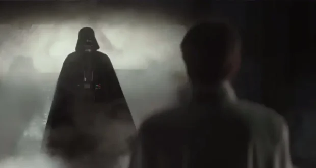 A dark, armored figure in a cape stands in a smoky, dimly lit corridor facing a person in a white uniform whose back is to the camera.