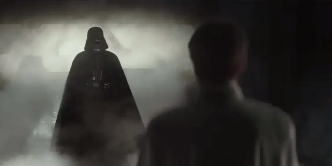 A dark, armored figure in a cape stands in a smoky, dimly lit corridor facing a person in a white uniform whose back is to the camera.
