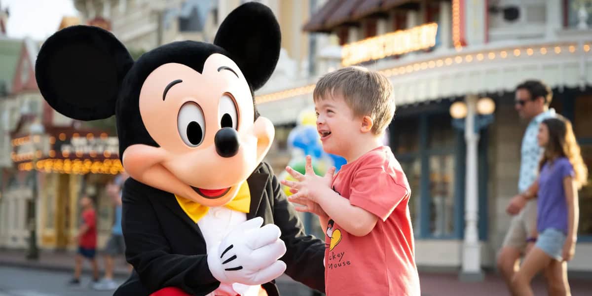 A young boy excitedly interacts with a person in a Mickey Mouse costume at an amusement park, with colorful buildings and people in the background.