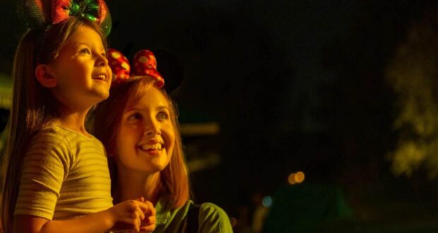 A young girl and a woman wearing Minnie Mouse ear headbands smile and look up, illuminated by warm light against a dark, nighttime background.