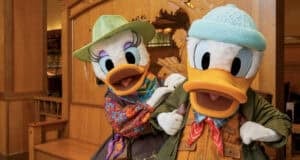 Donald Duck and Daisy Duck wear colorful outdoor adventure outfits and hats, standing together indoors, smiling and posing for the camera in a warmly lit wooden setting.