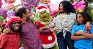 Four people smile and laugh as they pose with a person in a Grinch costume wearing a Santa suit, surrounded by colorful, whimsical decorations and large pink bows.
