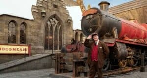 A train conductor in a red uniform stands in front of the Hogwarts Express steam train at Hogsmeade station, with stone buildings and a sign reading “Hogsmeade” in the background.