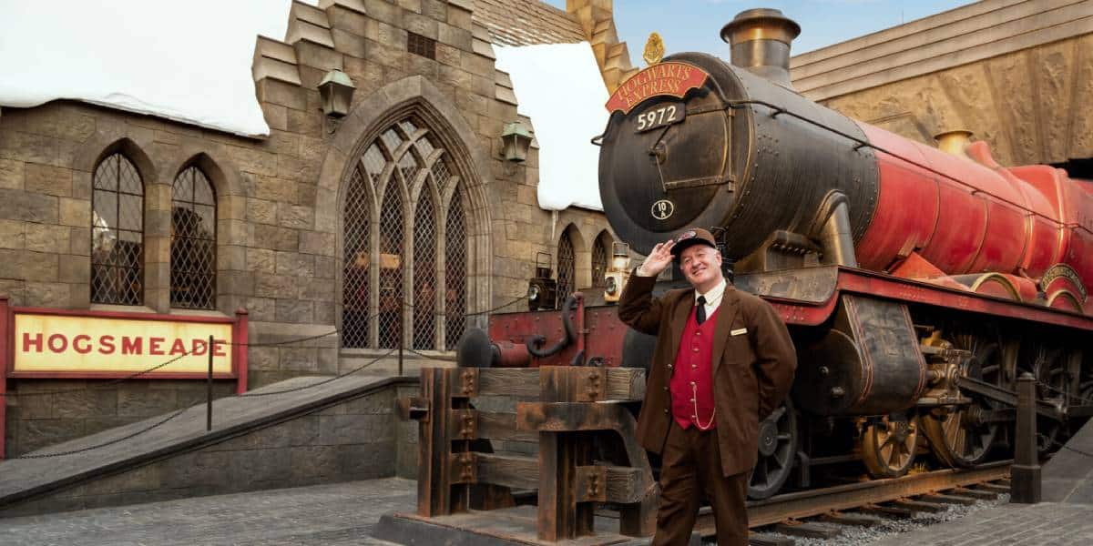 A train conductor in a red uniform stands in front of the Hogwarts Express steam train at Hogsmeade station, with stone buildings and a sign reading “Hogsmeade” in the background.