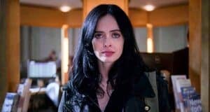 A woman with long black hair and a serious expression stands indoors, wearing a dark jacket. The background shows bookshelves and a softly lit room, suggesting a library or bookstore setting.