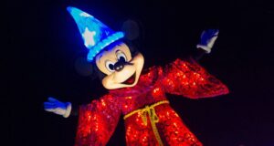 Mickey Mouse wears a glowing blue wizard hat and a sparkling red robe, arms outstretched and smiling brightly against a dark background.