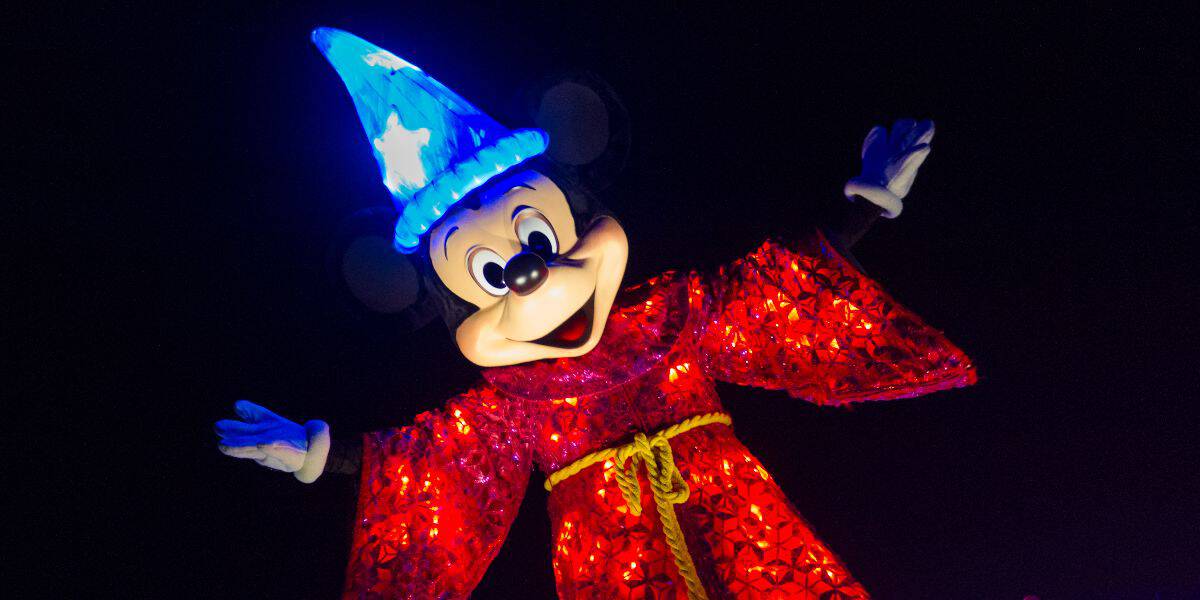 Mickey Mouse wears a glowing blue wizard hat and a sparkling red robe, arms outstretched and smiling brightly against a dark background.