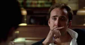 A man with light skin and brown hair sits indoors, resting his hand on his mouth and looking thoughtfully ahead. The background is blurred, suggesting a dimly lit room with lamps and shelves.