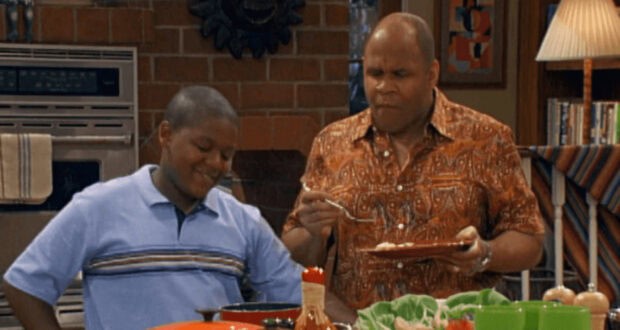 A man in a patterned shirt holds a plate and eats with a spoon, standing beside a smiling boy in a blue polo shirt in a cozy kitchen setting with vegetables and condiments on the countertop.