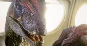 A close-up of a dinosaur with sharp teeth sitting inside an airplane, next to a window, under bright light.