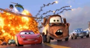 Animated cars with expressive faces speed down a road as a large explosion occurs behind them; a jet flies overhead with smoke trails. The scene is action-packed and colorful, set in the "Cars" movie universe.
