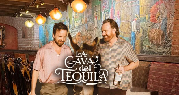 Two smiling men stand next to a donkey inside a rustic, colorful restaurant with a mural on the wall. Both are casually dressed, and one holds a bottle. The words “La Cava del Tequila” appear in front of them.