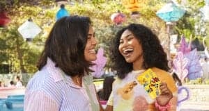 Two women laughing together outdoors at a colorful amusement park; one woman is holding a yellow ticket with Disney characters on it. Lanterns and trees are visible in the background.
