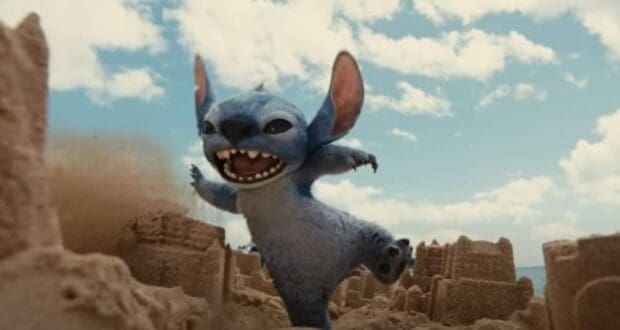 A blue, alien creature with large ears kicks over sandcastles on a beach, smiling mischievously under a partly cloudy sky—echoing the playful energy that early reviews for Lilo and Stitch suggest could make it a $1 billion film for Disney.