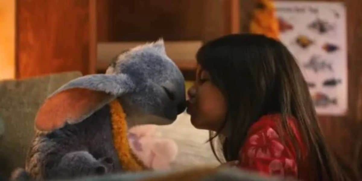 A young girl with long dark hair kisses the nose of Stitch, a blue alien character, in a cozy indoor setting. Stitch wears an orange lei, and the background features a poster with colorful illustrations.