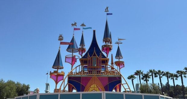 Colorful whimsical castle structure with blue and pink pointed towers, golden accents, and flags, set against a clear blue sky and surrounded by palm trees and greenery.