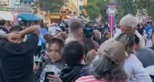 A crowded street scene at Disneyland, with people walking closely together, colorful banners, and illuminated buildings in the background. Trees line the street and park decorations are visible.