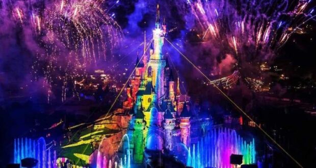 A brightly lit castle with rainbow-colored lights, surrounded by colorful fountains and fireworks bursting in the night sky above.