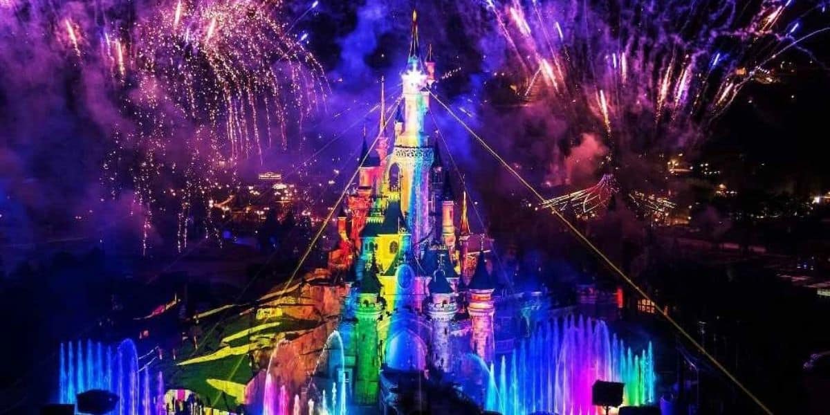 A brightly lit castle with rainbow-colored lights, surrounded by colorful fountains and fireworks bursting in the night sky above.