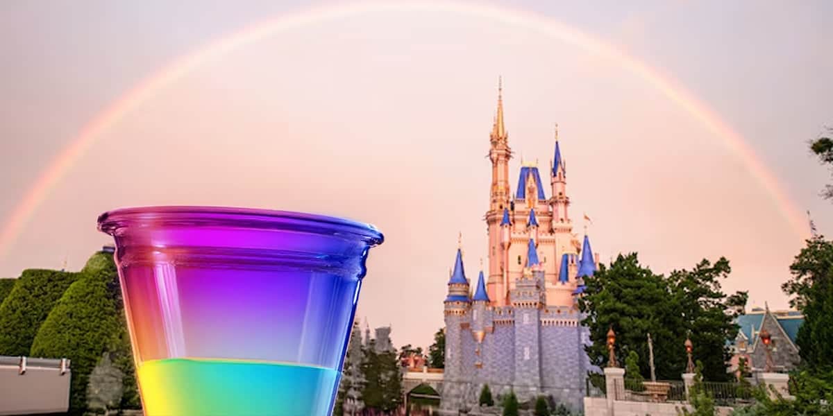 A colorful layered drink in a clear cup is in the foreground, with a fairytale castle and a vivid rainbow arching across a pastel sky in the background. Lush greenery surrounds the castle.