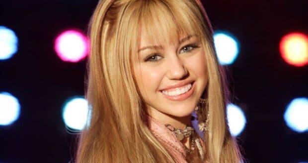 A young woman with long, straight blonde hair and bangs smiles at the camera. She is wearing a pink outfit, and colorful lights glow in the blurry background.