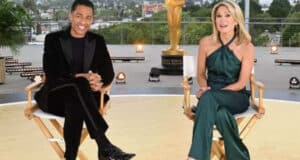 Amy Robach and TJ Holmes are seated on director's chairs on an outdoor terrace, with city buildings in the background and an Oscar statue behind them. TJ is wearing a black suit, while Amy stuns in a green jumpsuit. Both are smiling and appear to be hosting an event.