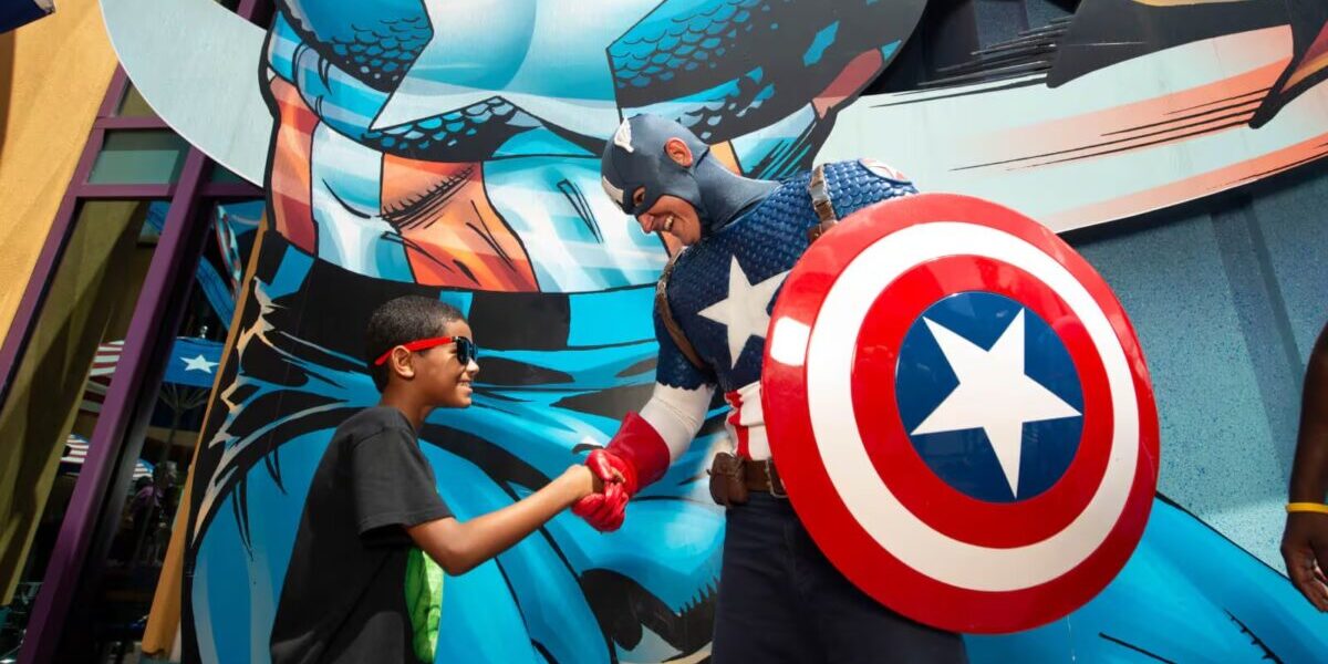 A smiling child wearing sunglasses shakes hands with a person dressed as Captain America, who holds a shield. They stand in front of a large Captain America mural.