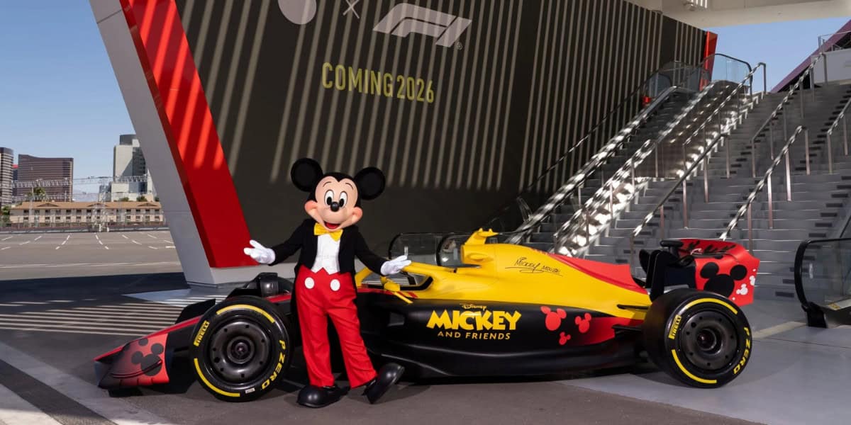 Mickey Mouse stands next to a yellow and black Formula 1 race car featuring Disney Mickey and Friends branding and graphics, with a sign in the background reading "COMING 2026 season.