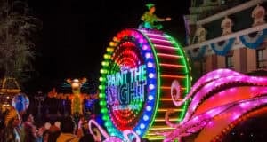 A vibrant nighttime Disneyland parade features a colorful, illuminated float with "Paint the Night" glowing brightly. A character is perched atop while neon figures dazzle in the background, captivating spectators watching from the sidelines or enjoying through a live stream.