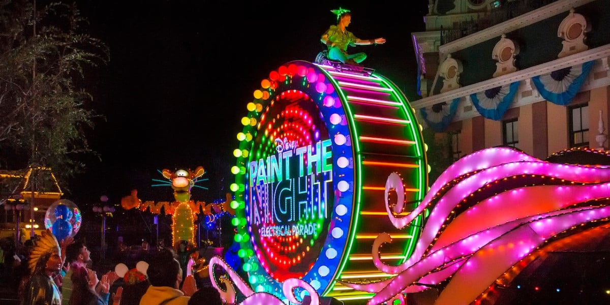 A vibrant nighttime Disneyland parade features a colorful, illuminated float with "Paint the Night" glowing brightly. A character is perched atop while neon figures dazzle in the background, captivating spectators watching from the sidelines or enjoying through a live stream.