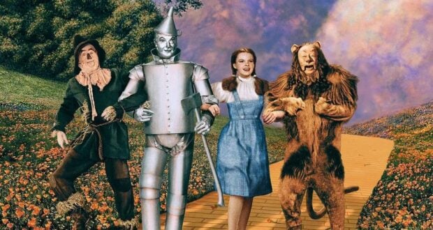 Four characters—the Scarecrow, Tin Man, Dorothy, and Cowardly Lion—walk arm in arm down a yellow brick road, surrounded by colorful flowers and greenery, from "The Wizard of Oz.