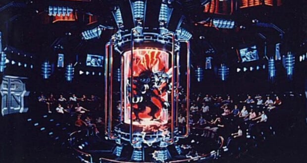 A futuristic auditorium with people seated in rows, watching a large glass chamber in the center that contains a menacing alien creature surrounded by glowing red and blue lights. Monitors and high-tech equipment fill the dark room.