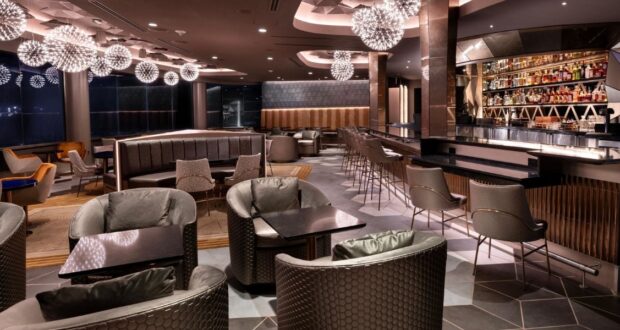 Modern lounge bar with stylish lighting, plush armchairs, dark tables, high chairs at a sleek bar, and shelves stocked with bottles. Warm, ambient lighting creates a cozy and inviting atmosphere.