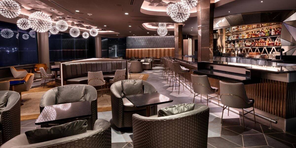 Modern lounge bar with stylish lighting, plush armchairs, dark tables, high chairs at a sleek bar, and shelves stocked with bottles. Warm, ambient lighting creates a cozy and inviting atmosphere.