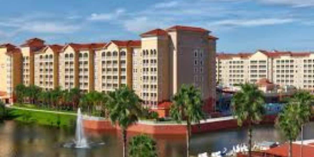 A large resort-style hotel with multiple stories, red-tiled roofs, and beige walls, surrounded by palm trees, a small lake with a fountain, and blue sky in the background.