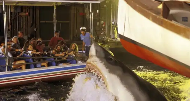 A mechanical shark emerges from the water near a boat, causing a group of tourists seated in a nearby tour boat to react with excitement and fear. The scene appears to be part of an amusement park attraction at Universal Orlando, with a staff member observing in the background.