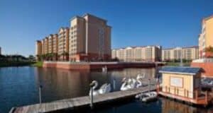 A dock with swan-shaped paddle boats extends over a calm lake, with several large multi-story apartment buildings and a clear blue sky in the background.