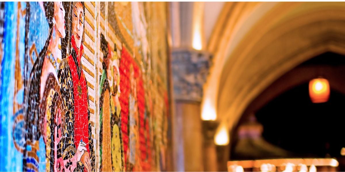A colorful mosaic mural on a wall, featuring people in historic attire—echoing the style seen in the Nazi behind Disney World and Disneyland murals—with an arched hallway and warm lighting blurred in the background.