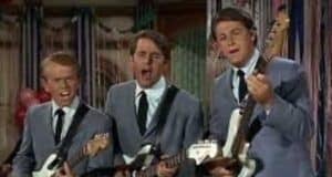 Three men wearing matching gray suits and black ties sing and play electric guitars together on stage, with colorful decorations in the background.