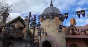 A whimsical building with a stone archway and turret, decorated with lanterns and purple banners featuring yellow sun symbols, set against a partly cloudy sky.