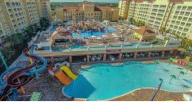 Aerial view of a large resort featuring multiple swimming pools, water slides, lounge chairs, and several tall yellow buildings surrounding the area on a sunny day.