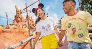 A smiling couple wearing Mickey Mouse-themed shirts and Minnie Mouse ears hold hands while walking at a Disney theme park, with Big Thunder Mountain Railroad and a bright blue sky in the background.
