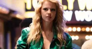 A woman with long blonde hair and hoop earrings wears a shiny green jacket and walks at night in front of bright, blurry, marquee lights and a partially visible "Hollywood" sign.