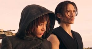 Two people stand together outdoors; one wears a hooded cloak with dreadlocks visible, and the other has long hair tied back, both looking serious and gazing into the distance under a warm light.