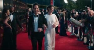 A man in a black tuxedo and a woman in a white dress, channeling Ant-Man-level confidence, walk arm-in-arm on a red carpet, surrounded by photographers, reporters, and other guests at a glamorous event.
