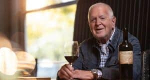 An older man sits at a table indoors, holding a glass of red wine and smiling. A bottle of wine is on the table next to him. Warm light comes through a window in the background.