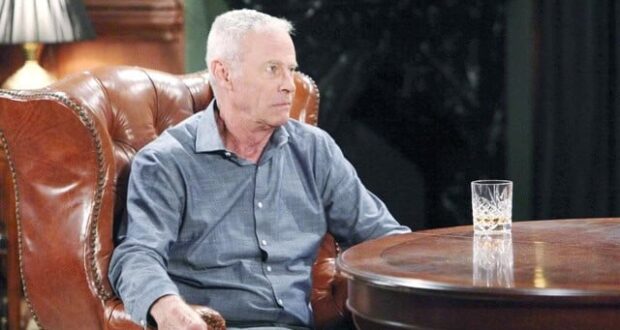 An older man with short gray hair sits in a brown leather armchair at a wooden table, wearing a blue shirt. A glass of whiskey is on the table in front of him. The background is dimly lit.