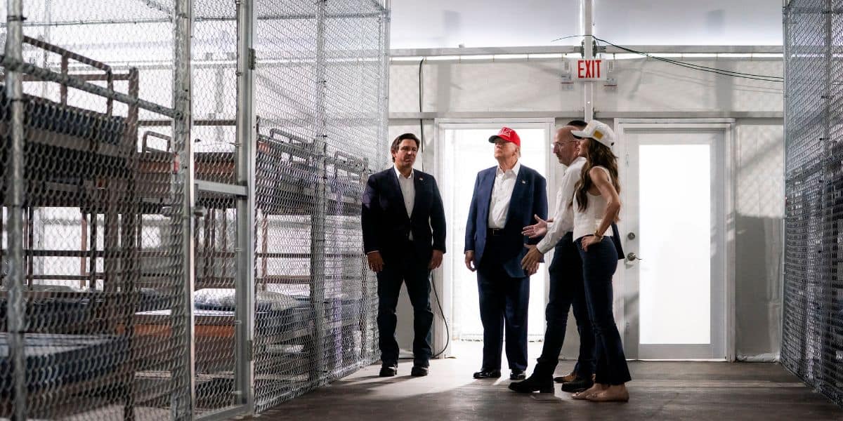 Four adults stand and talk inside a facility with metal bunk beds and chain-link fencing, near a door marked with an exit sign. Sunlight shines through windows above the beds.