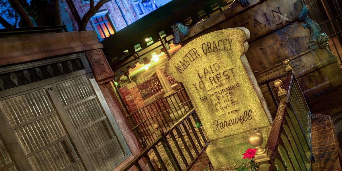 A gravestone inscribed “Master Gracey Laid To Rest” stands in front of an iron gate at night, surrounded by gold fencing and red flowers, with spooky decorations and dim lighting setting an eerie mood.